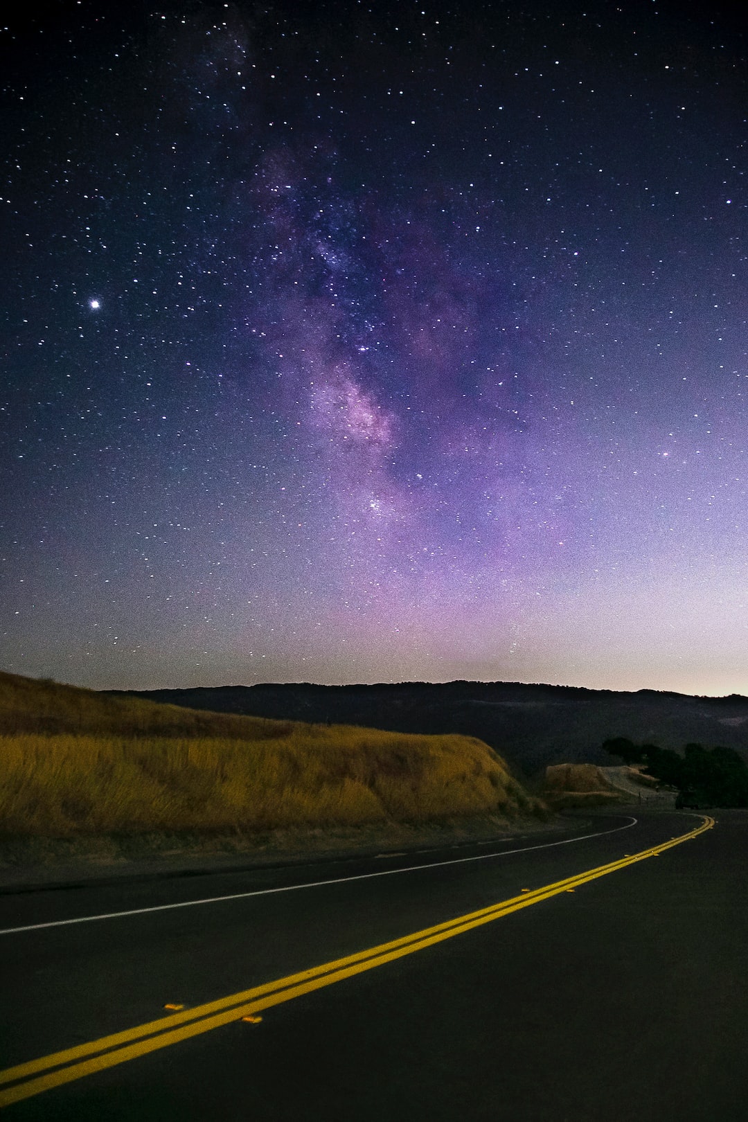 A road winding around grassy hills with a view of the Milky Way and stars in the sky in Del Valle near Livermore, California.