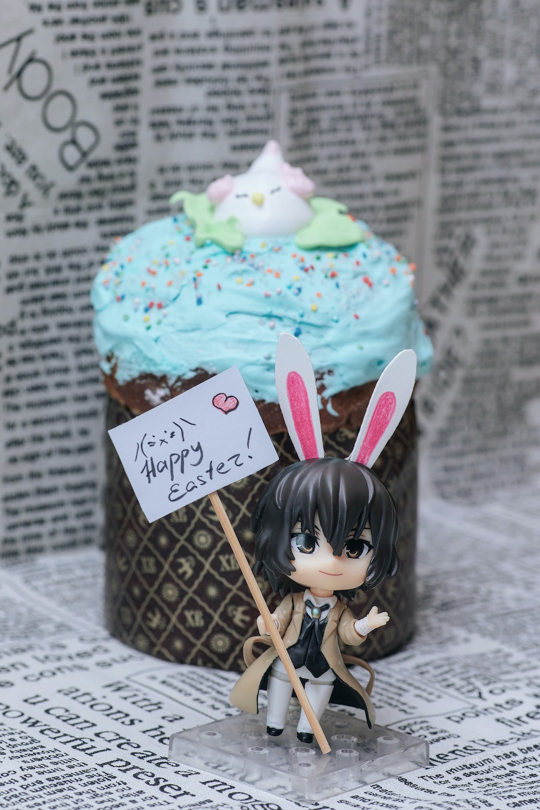 Anime character with a sign in his hand. The sign says "Happy Easter" with a heart on its side! In the background - newspaper headlines and kulich. The background is blurred, only individual letters can be seen.