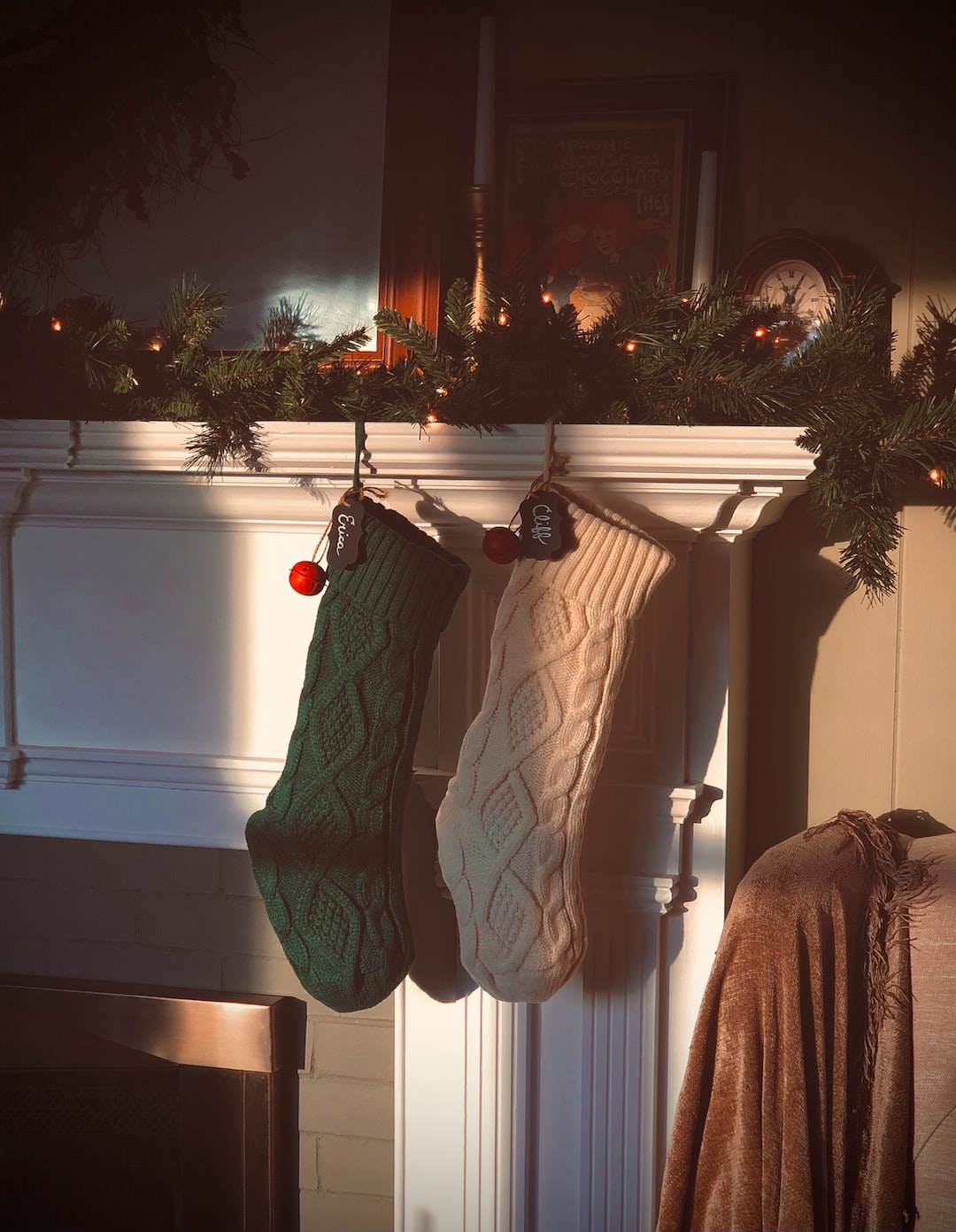 A green knitted holiday stocking and a white knitted holiday stocking sitting on an ornate white fireplace mantle as sun pours through a window and highlights the stockings. A red jingle bell and name tags sit on the stockings.