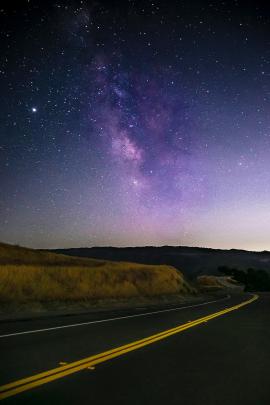 A road winding around grassy hills with a view of the Milky Way and stars in the sky in Del Valle near Livermore, California.