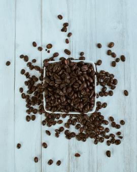 Square white bowl full of brown coffee beans spilling onto white wooden table