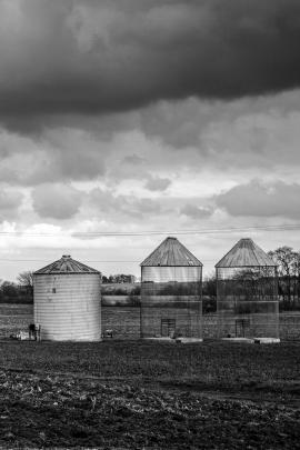 Three grain bins in rural country during afternoon on overcast cloudy day.