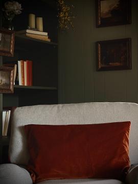 A close up of a dark, cozy reading corner. Antique bronze framed art prints sit on the walls and bookshelf. An off white colored English armchair looks inviting, ready for anyone to curl up with a good book.