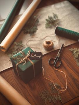 A newly wrapped Christmas present sitting on top of a wooden table with green wrapping paper and brown wrapping paper rolls surrounding the gift. Winter tree sprigs surround the wrapped present along with some twine and old scissors.