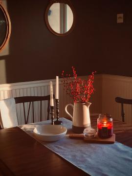 Red berries in a ceramic pitcher atop a vintage wooden table with black antique chairs. A Christmas candle burns on a wooden cutting board next to the pitcher along with black taper candle holders and an empty white bowl.