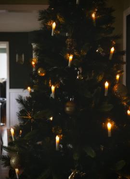 An up-close photo of a Christmas tree adorned with candle shaped lights and miscellaneous gold ornaments. The background includes dark green walls with golden wall sconces.
