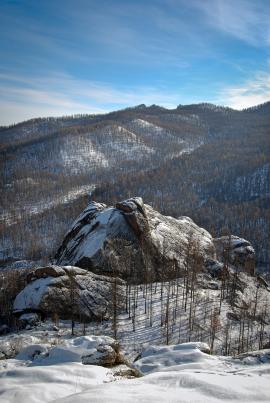 Place only accessible on horseback, picture of a lone boulder covered with snow with dense forest mountains in the backdrop. 