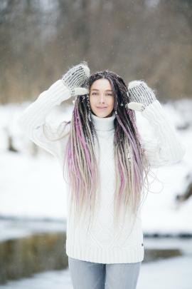Snowy photo shoot with a sensual young woman in a white sweater. Beautiful girl with long colored dreadlocks posing against the river during a snowfall
