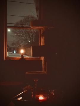 A glass jar candle burning on a dark winter night. A fake holiday candle is shining brightly in a window that displays a quiet, snowy Christmas scene in the background.